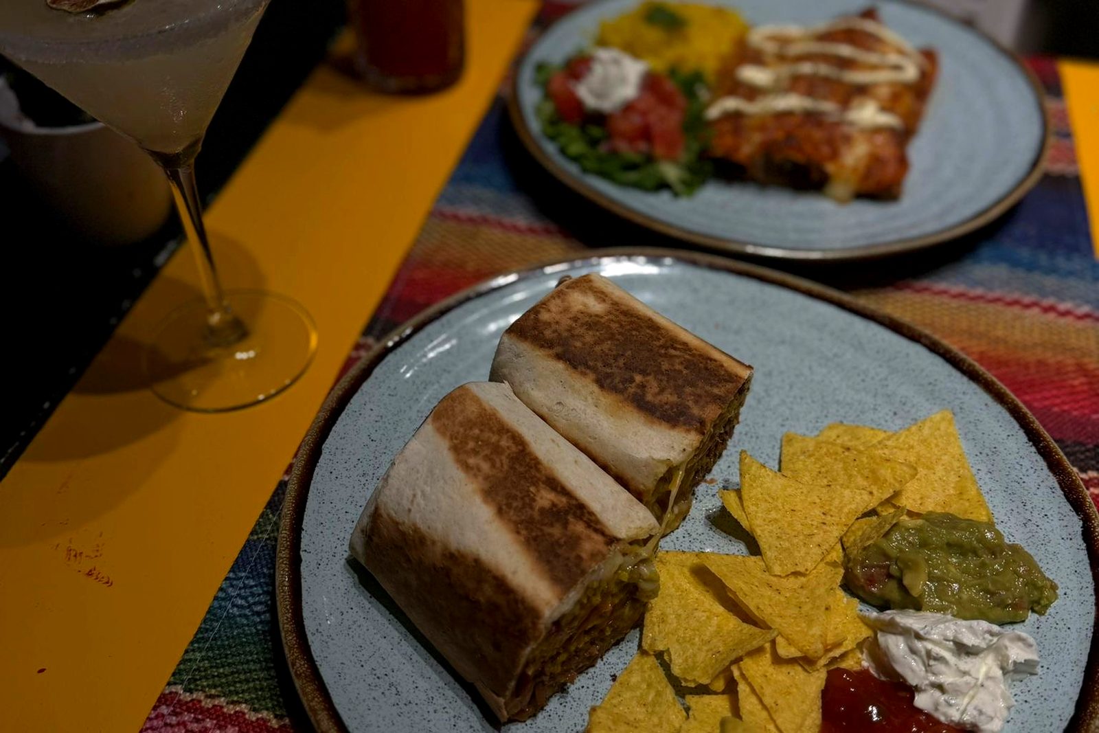 A plate of burrito halves with nachos, guacamole, sour cream and salsa, with a frozen margarita beside it
